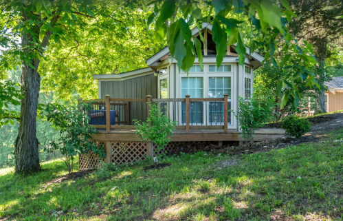 A small, charming cabin with a deck, surrounded by greenery and trees in a sunny outdoor setting.