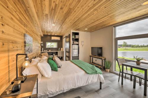 Cozy bedroom with wooden ceiling, large window, and modern decor, featuring a bed, kitchenette, and dining table.