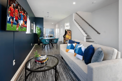 Modern living room with a sofa, coffee table, and a TV showing a soccer game, featuring a dining area and staircase.