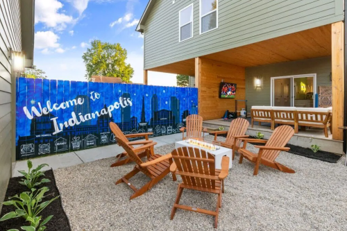 A cozy outdoor seating area with Adirondack chairs, a fire pit, and a colorful mural reading "Welcome to Indianapolis."