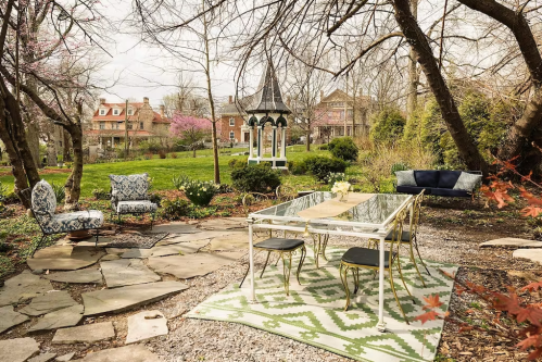 A serene garden with a glass table, patterned rug, and seating surrounded by trees and blooming flowers.