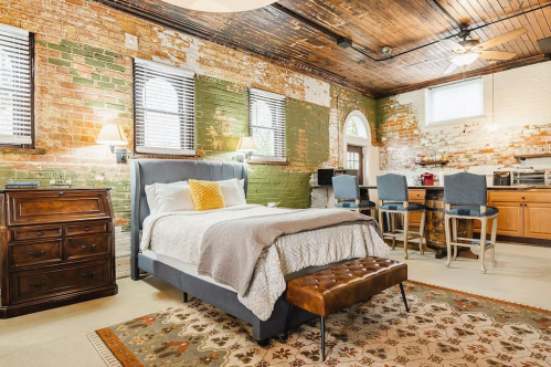 Cozy bedroom with a gray bed, vintage decor, exposed brick walls, and a small kitchen area in a stylish loft.