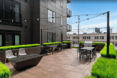 Rooftop patio with seating, grill, and string lights, surrounded by greenery and city skyline in the background.