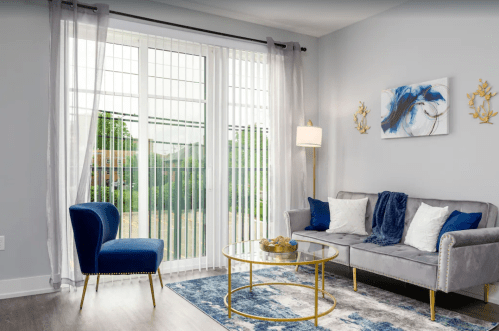 A modern living room with a gray sofa, blue chair, gold coffee table, and large windows with sheer curtains.