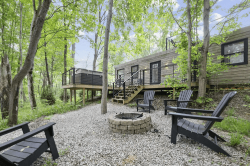 A modern cabin surrounded by trees, featuring a fire pit and outdoor seating area on a gravel path.