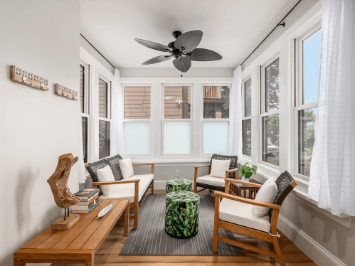 Bright sunroom with large windows, comfortable seating, a ceiling fan, and a green patterned ottoman.
