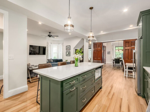 Modern kitchen with green cabinetry, a large island, pendant lights, and an open layout leading to a living area.