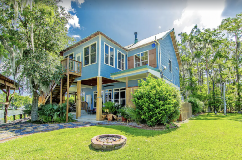 A blue two-story house surrounded by greenery, featuring a fire pit and a view of the water.