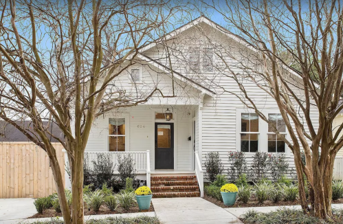 A charming white house with a front porch, surrounded by trees and colorful potted plants.