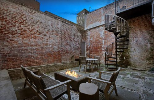 A cozy outdoor courtyard with a fire pit, seating, and a spiral staircase against a brick wall backdrop.