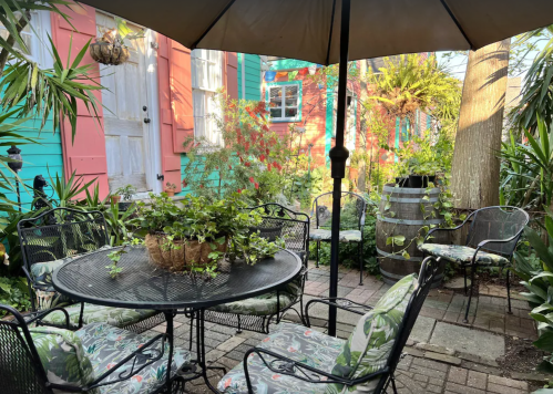 A cozy outdoor patio with a round table, chairs, and lush greenery, surrounded by colorful buildings and plants.