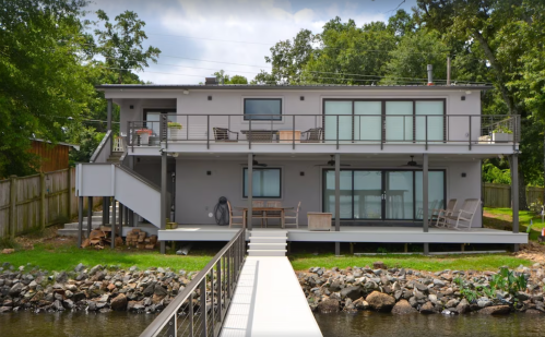 A modern two-story house by a lake, featuring large windows, a deck, and a wooden walkway leading to the water.