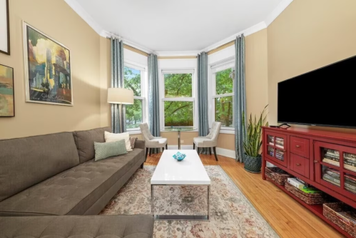 Cozy living room with a gray sofa, coffee table, and two chairs by large windows, featuring warm tones and decorative plants.