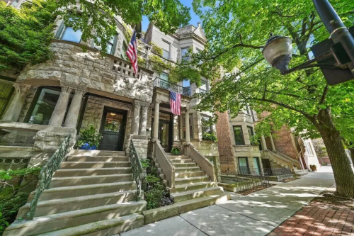 Historic stone buildings with American flags, surrounded by lush green trees and a sunny blue sky.