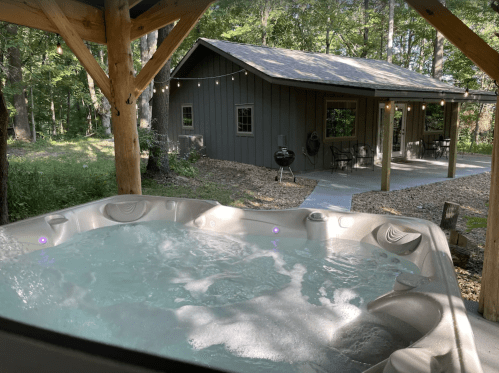 A hot tub with bubbling water in the foreground, a cozy cabin surrounded by trees in the background.