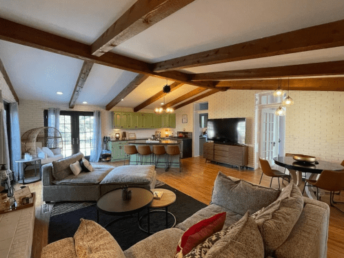 Spacious living area with wooden beams, a cozy sofa, kitchen with green cabinets, and dining space. Bright and inviting.