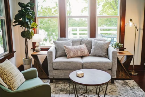 Cozy living room with a gray sofa, green chair, round coffee table, and large windows overlooking greenery.