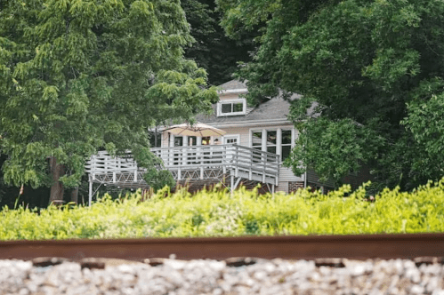 A house with a deck nestled among trees, viewed from a distance near a railway track.