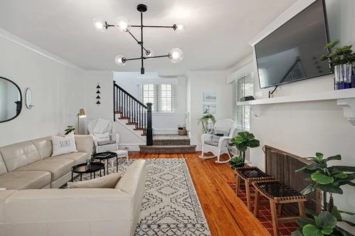 Modern living room with a light fixture, beige sofas, a patterned rug, plants, and a staircase in the background.