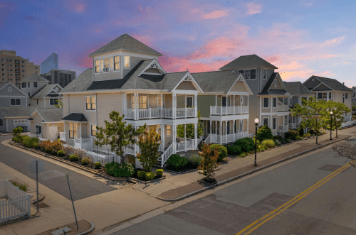A charming multi-story house with a wraparound porch, surrounded by greenery, at sunset in a quiet neighborhood.