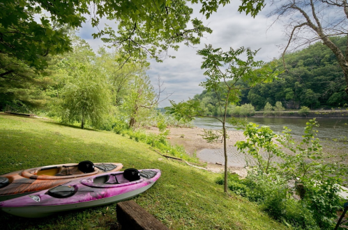 Two kayaks rest on a grassy bank by a calm river, surrounded by lush greenery and a cloudy sky.