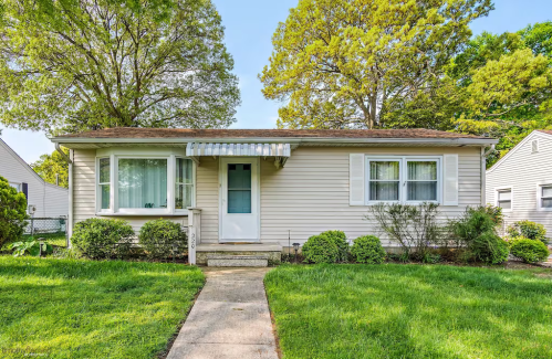 A single-story house with a front porch, surrounded by green grass and trees, under a clear blue sky.