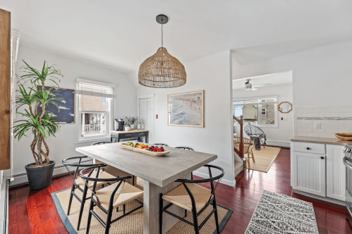 Bright dining area with a wooden table, woven pendant light, and a fruit centerpiece, surrounded by plants and natural light.
