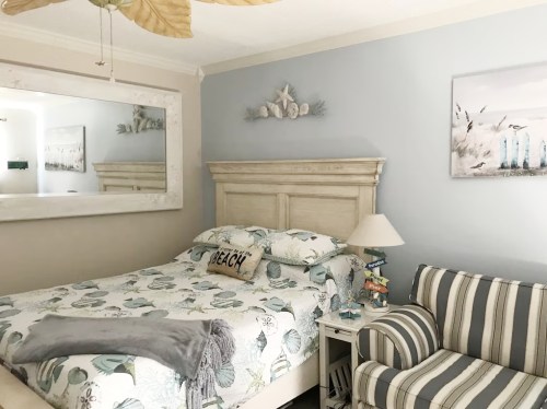Cozy bedroom with a beach theme, featuring a bed, striped chair, and decorative mirror on the wall.