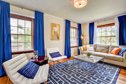 Cozy living room with blue curtains, a beige sofa, modern chairs, and a geometric rug, featuring natural light from windows.