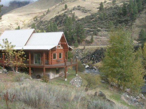 A cozy log cabin by a river, surrounded by rolling hills and trees under a cloudy sky.