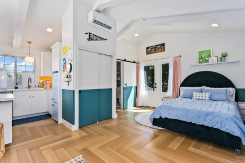 Cozy bedroom with a blue bedspread, wooden flooring, and a small kitchen area, featuring bright natural light.