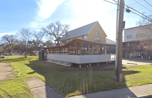A two-story building with a wooden deck, surrounded by grass and trees, on a sunny day.