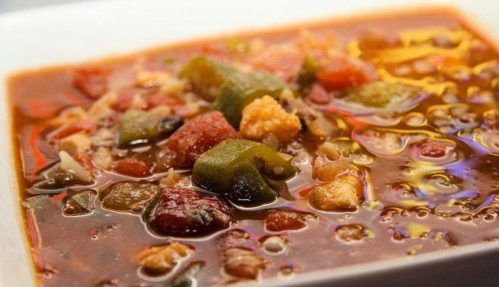 A close-up of a hearty vegetable soup with colorful ingredients like okra, tomatoes, and spices in a white bowl.