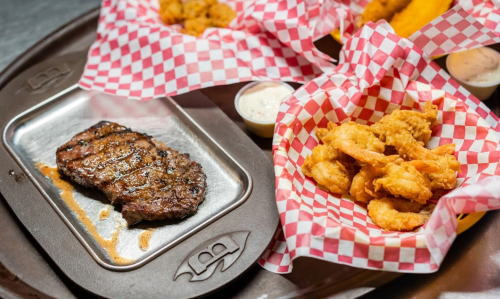 A metal tray with grilled steak beside baskets of fried shrimp and dipping sauces, all lined with red-checkered paper.
