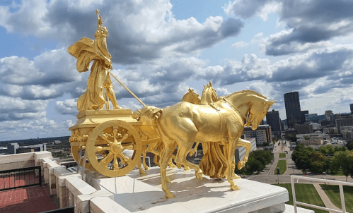 Golden statue of a figure in a chariot pulled by horses, set against a cloudy sky and city skyline.