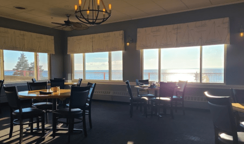 Bright restaurant interior with wooden tables and chairs, large windows overlooking a scenic view of the ocean.