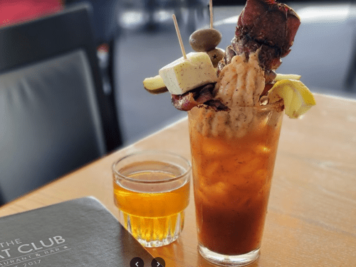 A tall cocktail garnished with shrimp, bacon, cheese, and olives, alongside a small glass of whiskey.