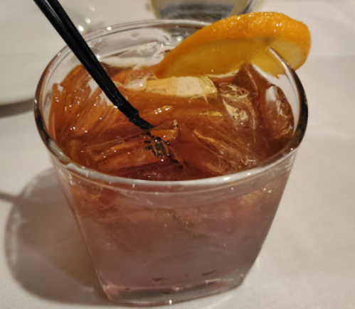 A close-up of a glass filled with a brown drink, ice, and a slice of orange, with a black straw.
