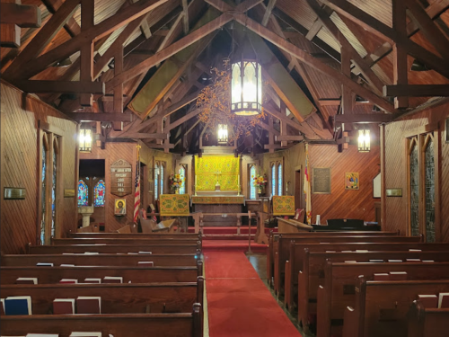 Interior of a rustic church with wooden beams, pews, and a red carpet leading to an altar adorned with flowers.