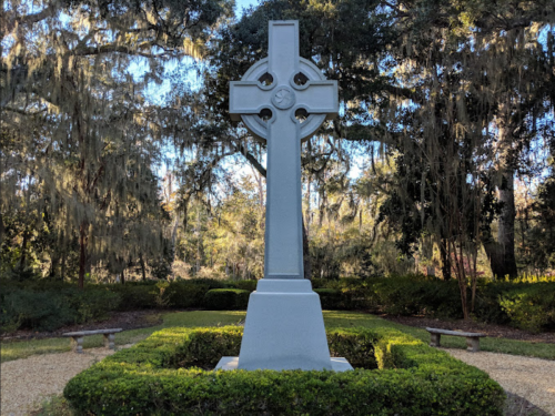 A tall white Celtic cross stands in a garden surrounded by greenery and benches, with trees in the background.