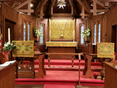 Interior of a church with wooden walls, colorful altar cloths, and floral arrangements, featuring a central altar.