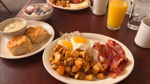 A hearty breakfast plate with eggs, bacon, potatoes, biscuits, and a glass of orange juice on a table.