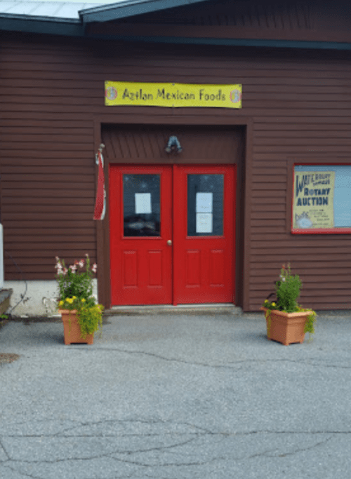 Red double doors of Aztlan Mexican Foods, flanked by potted plants, with a sign for a rotary auction nearby.
