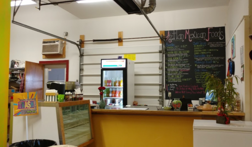 Interior of a Mexican food shop featuring a counter, menu board, and a refrigerator with drinks. Brightly colored decor.