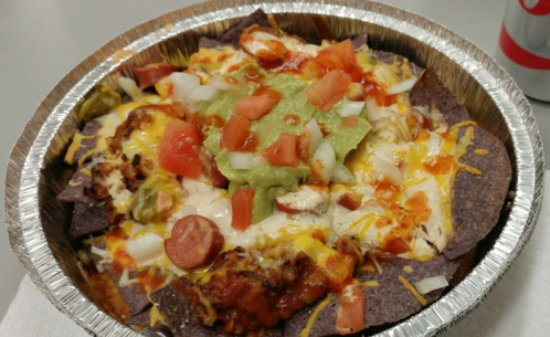 A bowl of nachos topped with cheese, guacamole, diced tomatoes, onions, and shredded chicken on blue corn chips.