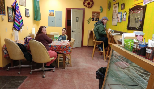 Two women sit at a table in a colorful café, while a man sits at the counter, surrounded by vibrant decor.