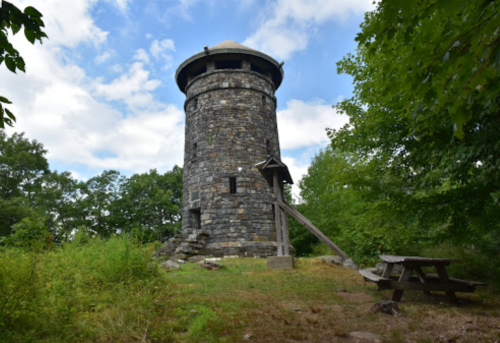 A stone tower surrounded by trees, with a picnic table in the foreground under a partly cloudy sky.