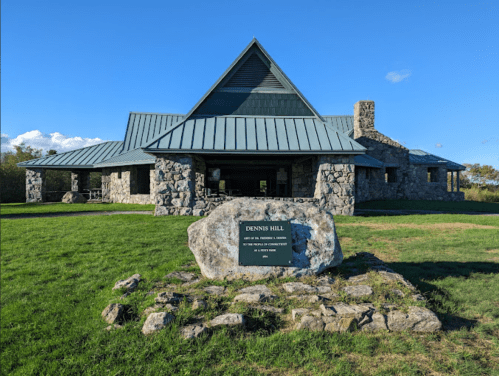 A stone building with a metal roof, surrounded by grass, featuring a sign that reads "Dennis Hill." Clear blue sky above.