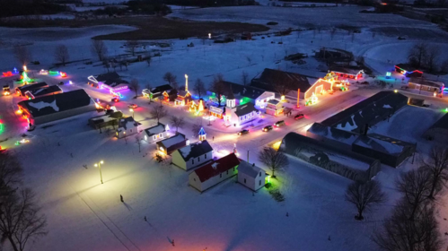 Aerial view of a snowy village illuminated with colorful holiday lights, showcasing festive decorations on buildings.