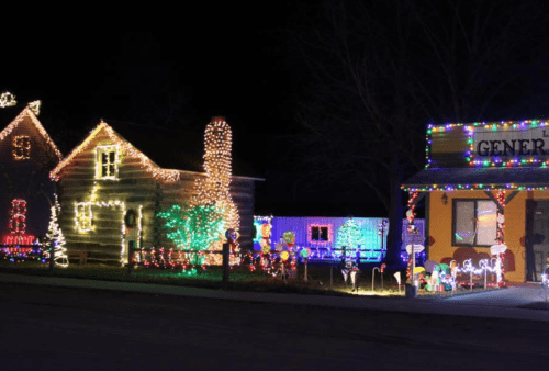 A festive scene of a log cabin and a general store, both adorned with colorful holiday lights at night.